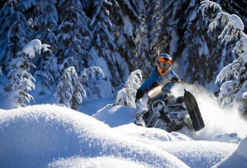 A person riding a snowmobile through deep snow in a forest of evergreen trees. Snow is flying around them.