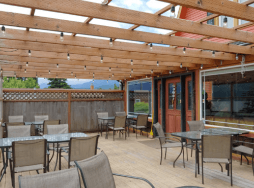 Outdoor dining area with wooden beams, string lights, and tables set on a wooden deck. Greenery in the background.