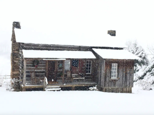 A rustic wooden cabin covered in snow, with a porch and an American flag visible through the falling snowflakes.