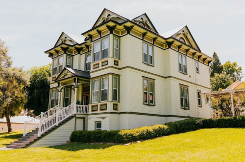 A large, historic two-story house with a green and yellow exterior, featuring ornate architectural details and a front porch.