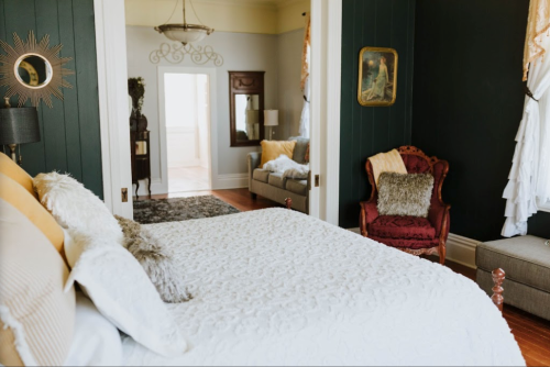 Cozy bedroom with a white quilt, dark green walls, and a view of a living area through an open doorway.