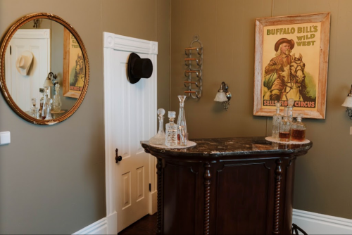 A vintage bar area with a mirror, hat, and a Buffalo Bill poster on the wall, featuring glassware and a marble countertop.