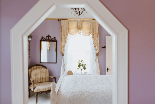 A cozy bedroom with a purple wall, a bed, a vintage chair, and a window adorned with elegant curtains.