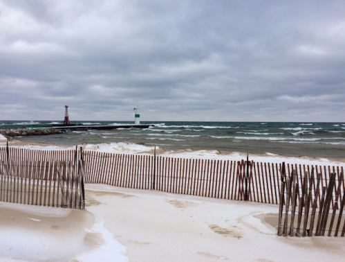 A beach scene with a wooden fence, waves, and a lighthouse in the distance under a cloudy sky.