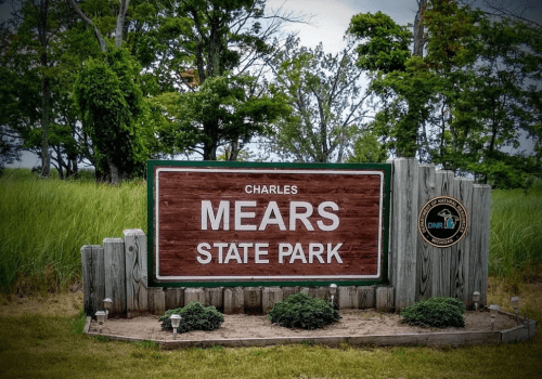 Sign for Charles Mears State Park, featuring wooden design and green accents, surrounded by grass and trees.