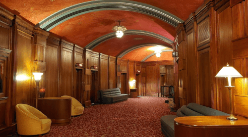 A vintage hallway with wooden paneling, red carpet, and soft lighting from lamps, featuring seating areas.