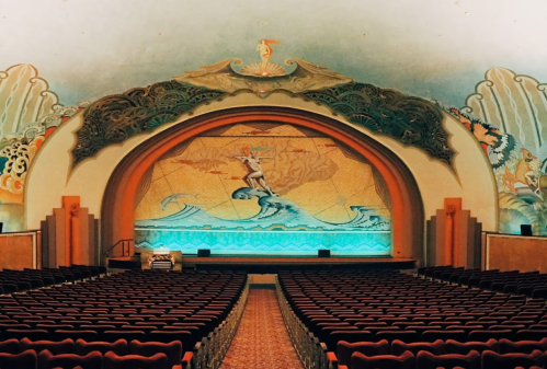 Interior of a vintage theater featuring a decorative stage with ocean-themed murals and rows of red seats.