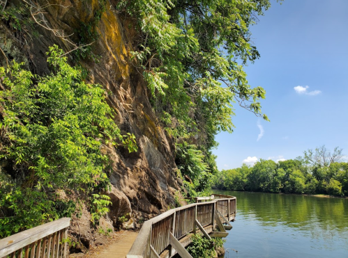 A wooden walkway curves along a river, flanked by lush greenery and a rocky cliff under a clear blue sky.