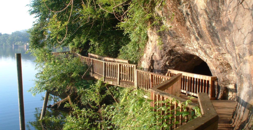 A wooden walkway leads to a cave by a calm river, surrounded by lush greenery and rocky cliffs.