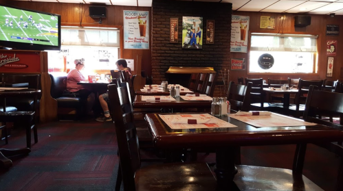 A cozy restaurant interior with wooden tables, a fireplace, and patrons enjoying their meals while watching a game on TV.