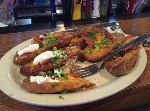 A plate of loaded potato skins topped with cheese, bacon, sour cream, and green onions, served at a bar.
