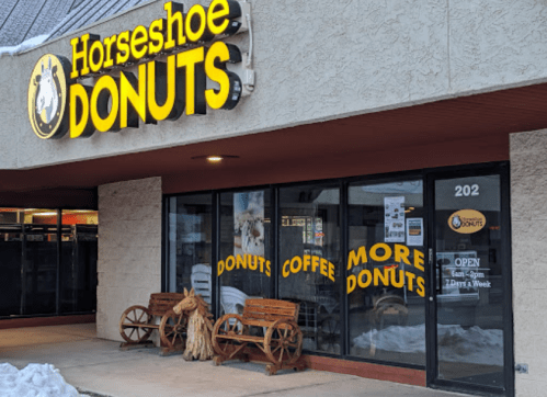 Exterior of Horseshoe Donuts shop featuring a sign, windows displaying donuts and coffee, and wooden benches outside.