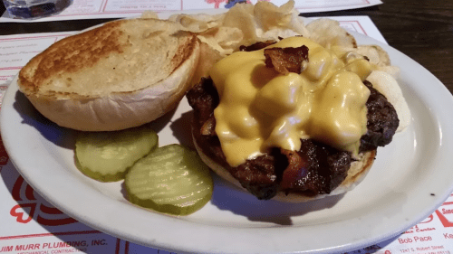 A juicy cheeseburger topped with bacon, served with pickles and potato chips on a plate.