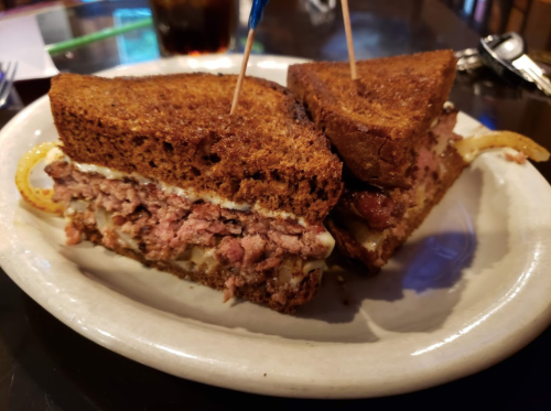 A close-up of a sandwich with dark rye bread, filled with meat and topped with mayonnaise, served on a white plate.