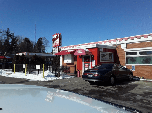 A red and white restaurant with a sign, surrounded by snow, featuring a parked car in front on a sunny day.
