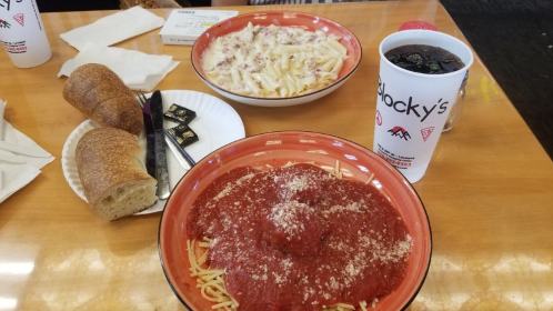 A table with two pasta dishes, bread rolls, and a drink in a cup, all set on a wooden surface.