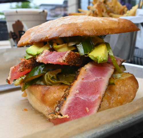 A close-up of a sandwich with seared tuna, avocado, and green peppers on a toasted bun, served on a tray.