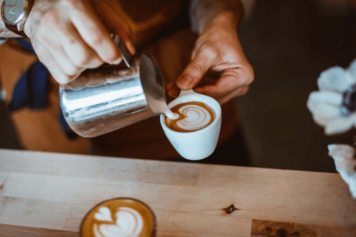 A barista pours steamed milk into a cup, creating a heart-shaped latte art design.