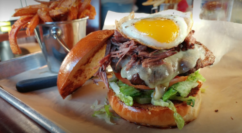 A juicy burger topped with melted cheese, pulled meat, lettuce, and a fried egg, served with sweet potato fries.
