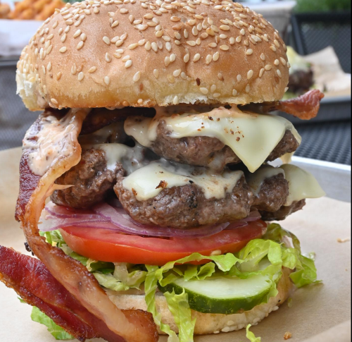 A towering burger with two beef patties, bacon, cheese, lettuce, tomato, and pickles on a sesame seed bun.