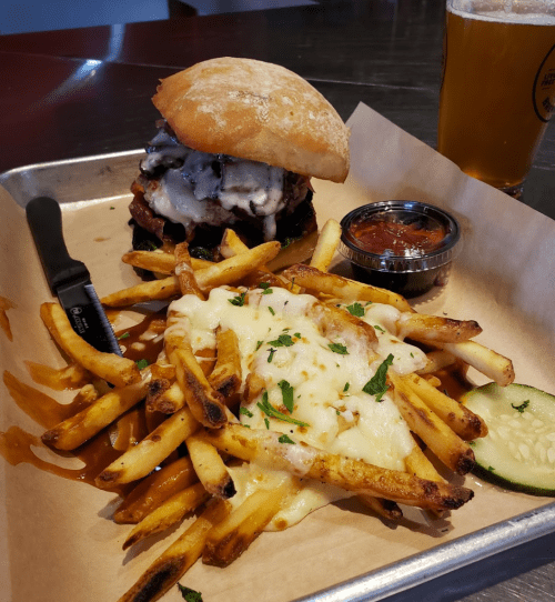A delicious burger with melted cheese on a bun, served with cheesy fries, a dipping sauce, and a pickle on a tray.
