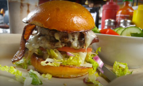 A close-up of a juicy burger with melted cheese, bacon, lettuce, and tomato on a toasted bun, served with fresh vegetables.