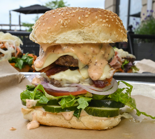 A towering burger with sesame seed bun, lettuce, tomato, pickles, cheese, bacon, and sauce, served on a paper tray.