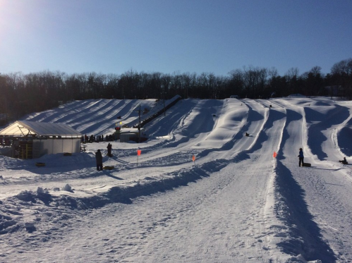 Snow-covered tubing hill with multiple lanes, a small building, and people enjoying the winter activity under a clear blue sky.