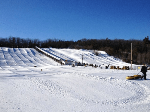 A snowy hill with multiple tubing lanes, people waiting in line, and a clear blue sky in the background.