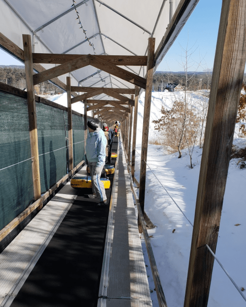 A snowy outdoor area with a covered conveyor belt for tubing, featuring people preparing to ride down the slope.