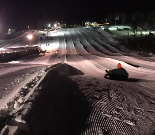 A person sledding down a snowy slope at night, with lights illuminating the hill and surrounding area.