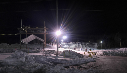 Nighttime scene of a snowy ski slope with illuminated jumps and skiers in action under bright lights.