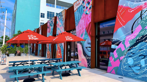 Colorful murals adorn a building's exterior, with red umbrellas over teal picnic tables in a vibrant outdoor space.