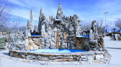 A rocky fountain surrounded by a paved area, with blue water and a clear blue sky in the background.