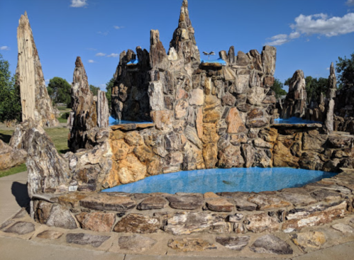 A rocky fountain structure with blue water, surrounded by natural stone formations and a clear blue sky.