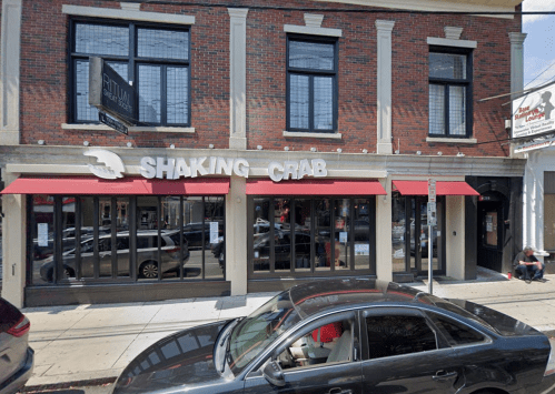 Exterior of Shaking Crab restaurant with large windows and red awnings, featuring a parked car in front.