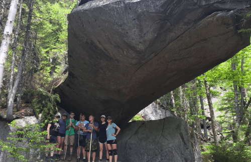 A group of six people stands under a large rock formation in a forested area, surrounded by trees and greenery.