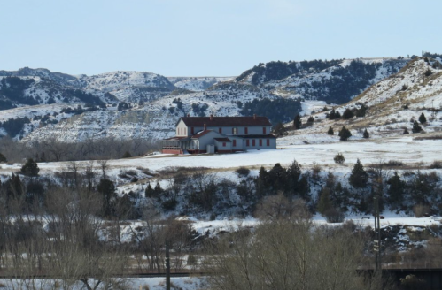 A snowy landscape featuring a large house on a hill, surrounded by rolling hills and sparse trees.