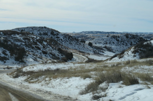 A winding road through snow-covered hills and grassy fields under a cloudy sky.