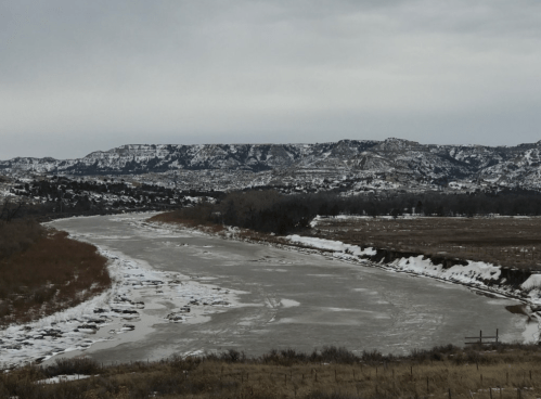 A frozen river winds through a snowy landscape, with mountains in the background under a cloudy sky.