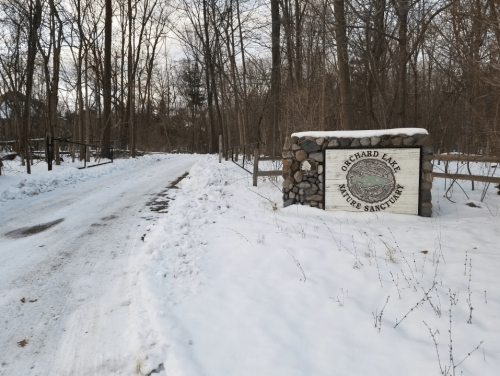 A snowy path leads to a stone sign for Orchard Lake Nature Sanctuary, surrounded by bare trees.