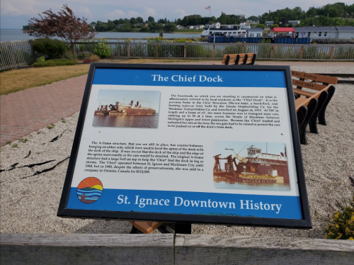 Sign about "The Chief Dock" in St. Ignace, detailing its history and significance, with a scenic waterfront view.