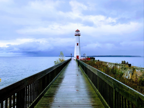 A wooden pier leads to a white lighthouse by the water, under a cloudy sky.