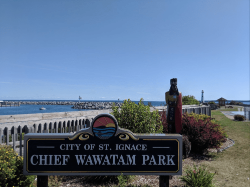 Sign for Chief Wawatam Park in St. Ignace, with a view of the waterfront and a statue in the background.