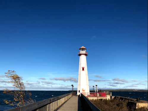 A white lighthouse with a red top stands on a pier, overlooking a blue ocean under a clear sky.