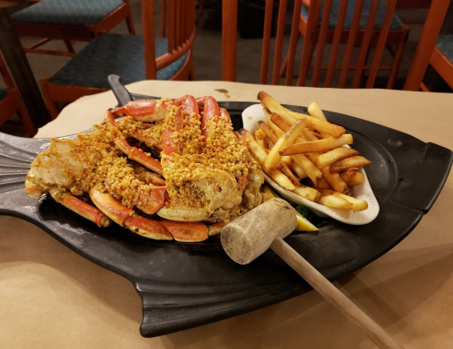 A platter of crab topped with breadcrumbs, served with a side of fries and a wooden mallet on a fish-shaped tray.