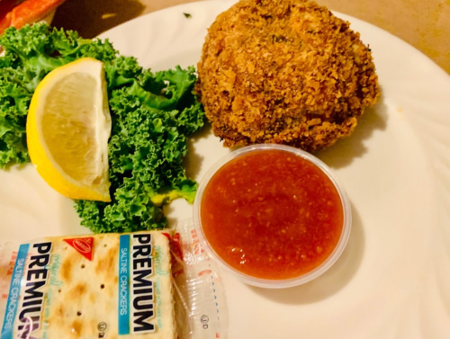 A plate with a golden-brown fried item, a lemon wedge, kale, a cup of red sauce, and a pack of saltine crackers.
