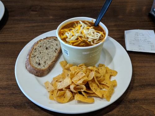 A bowl of chili topped with cheese, served with a slice of bread and a side of Fritos on a white plate.