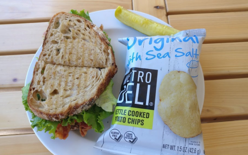 A plate with a sandwich made of lettuce and bacon, alongside a bag of kettle-cooked potato chips and a pickle.