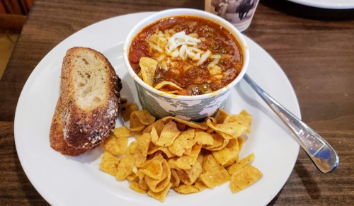 A bowl of chili topped with cheese, served with a side of Fritos and a slice of bread on a white plate.